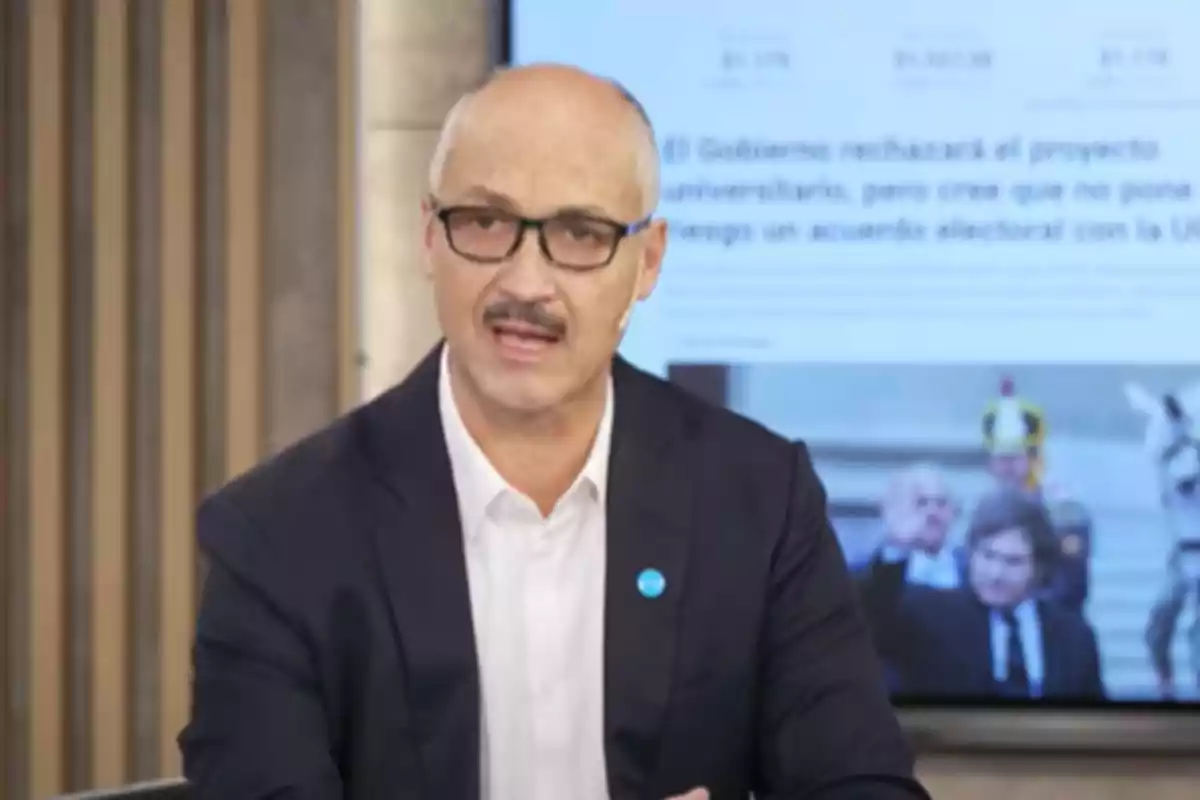 Hombre calvo con gafas y bigote hablando en un estudio de televisión con una pantalla de fondo mostrando texto y una imagen borrosa. Hombre calvo con gafas y bigote hablando en un estudio de televisión con una pantalla de fondo mostrando texto y una imagen borrosa.