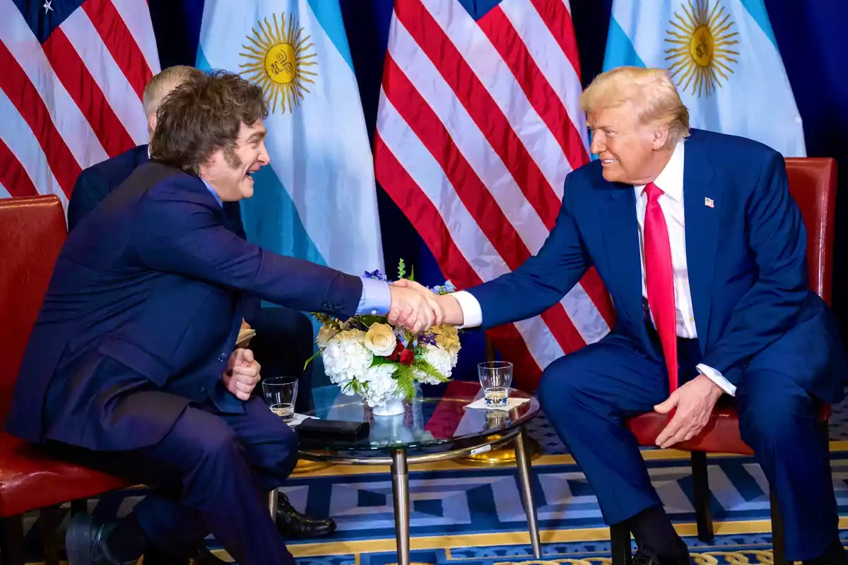 Dos personas estrechándose la mano mientras están sentadas frente a una mesa con banderas de Estados Unidos y Argentina de fondo. Dos personas estrechándose la mano mientras están sentadas frente a una mesa con banderas de Estados Unidos y Argentina de fondo.