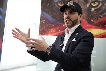 Abelardo de la Espriella con barba, traje, gorra de tigre y pin de Colombia, gesticula con ambas manos frente a un fondo con un tigre y la bandera